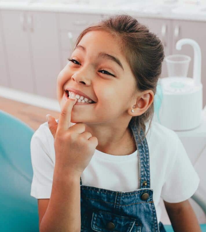 Smiling-girl-patient-child-teeth-healthy-smile-dentist A girl smiling pointing to her beautiful white teeth